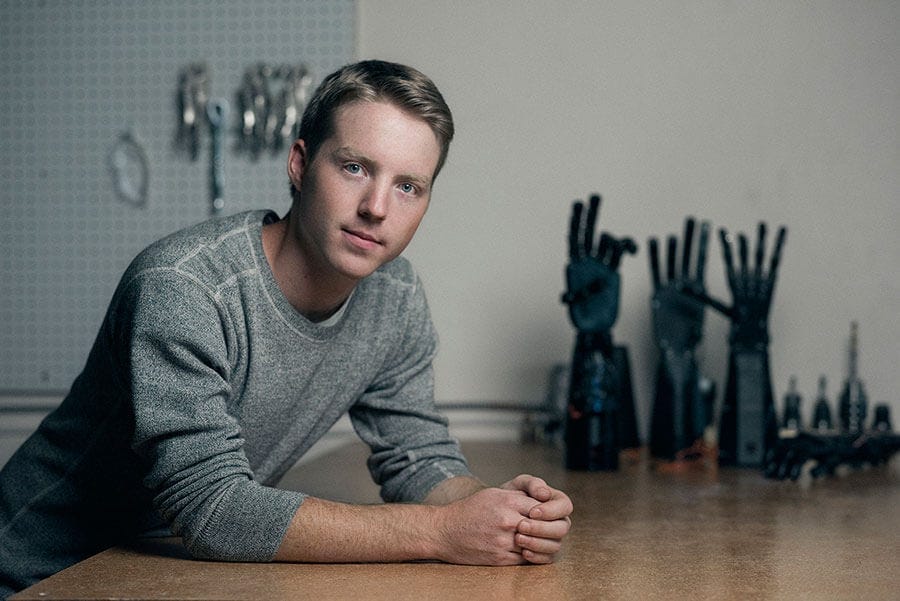 Easton LaChappelle leaning against a desk alongside several of his prosthetic inventions from Unlimited Tomorrow