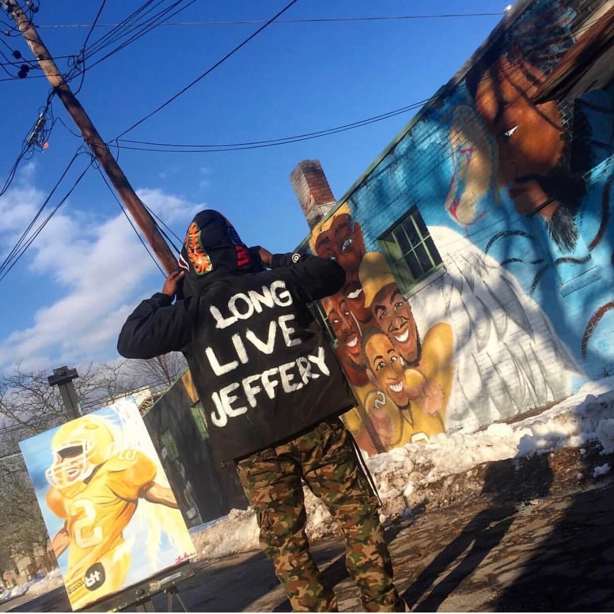 Jalani Lion standing in front of his Kingston mural wearing a jacket with the words "Long Live Jeffrey" on the back