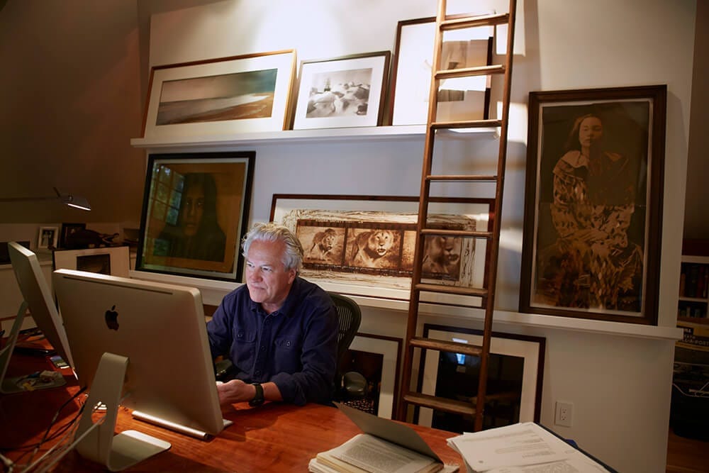 Jon Bowermaster working at a computer in an office in front of numerous art prints and photographs