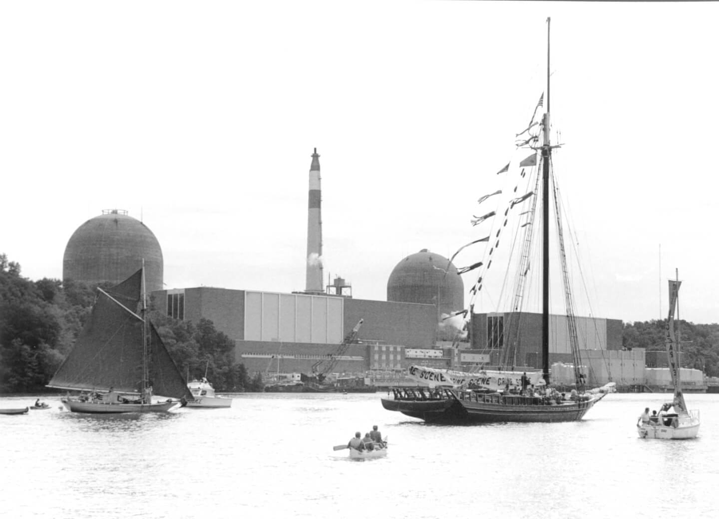 A black-and-white photo of the Sloop Clearwater and other boats in front of the Indian Point nuclear station on the Hudson River